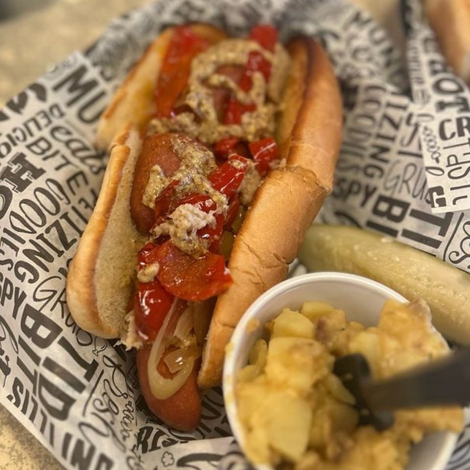 German hot dog with peppers, onions, and mustard, served with potato salad and a pickle.