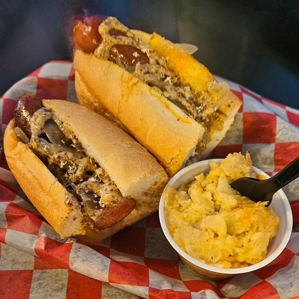 Two German bratwursts with mustard and onions, served with a side of mac and cheese.