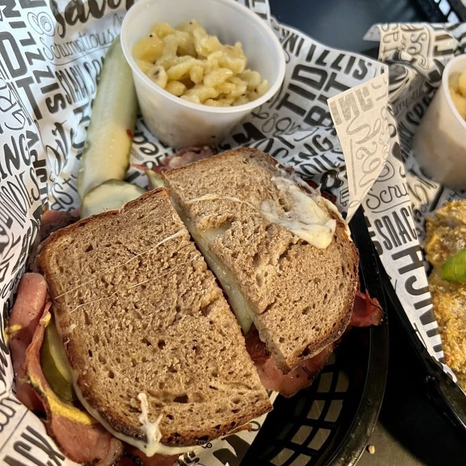 A German pastrami sandwich with melted cheese, pickles, and a side of spaetzle in a basket.
